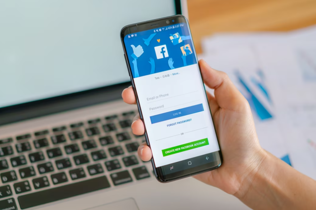 hand holding a smartphone with a Facebook platform on it in front of the opened laptop