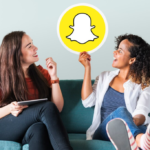 two women sitting on a green sofa, one holds a Snapchat picture and looks at it, other holds a tablet and looks at the picture
