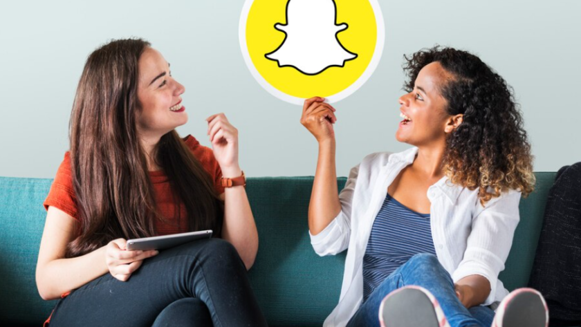 two women sitting on a green sofa, one holds a Snapchat picture and looks at it, other holds a tablet and looks at the picture