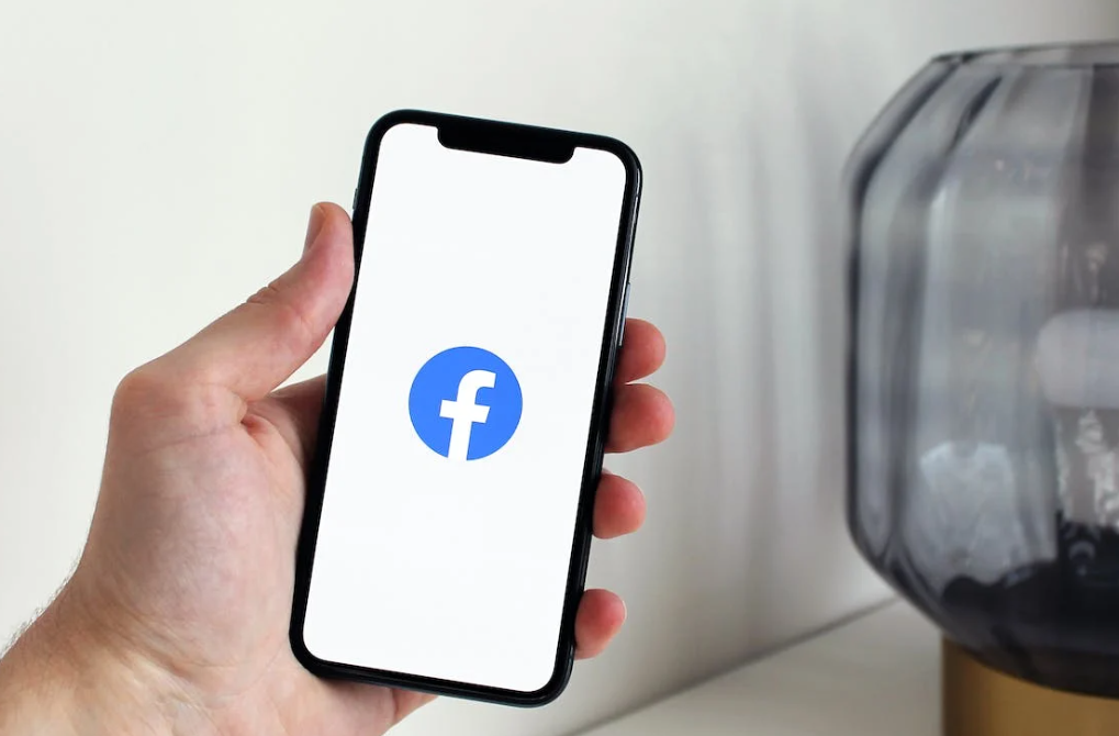 hand holding a phone with facebook icon on it, glass lamp in the corner